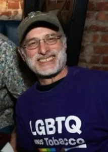 A smiling man with glasses and a beard wears a purple "LGBTQ minus Tobacco" shirt and a cap, sitting in a cozy room with brick walls.