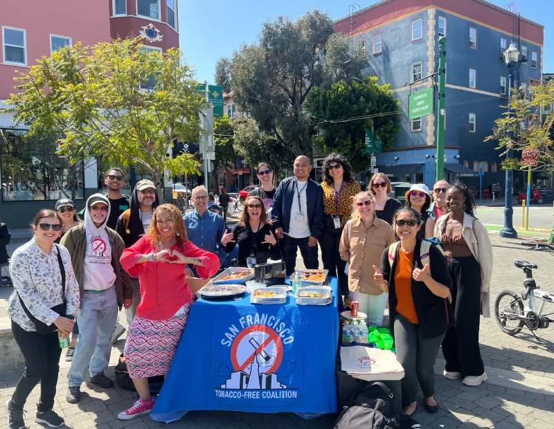 A diverse group of people smiling and standing around a table with food. The table is covered with a San Francisco Tobacco-Free Coalition cloth. Bright, sunny day in a lively urban setting.