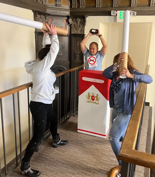 Three people are playfully using giant cigarette props; one is in a cigarette box costume with a no-smoking symbol. The scene is light-hearted and humorous.