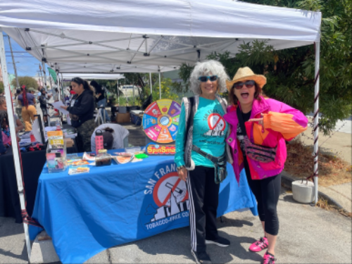 A smiling duo stands in front of a booth with a spinning wheel and a blue tablecloth displaying "San Francisco Tobacco-Free." They're under a white canopy at an outdoor event, conveying joy and activism.