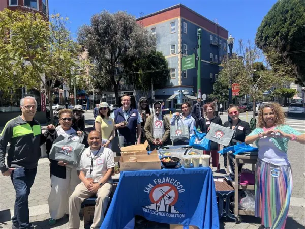 A group of smiling people at an outdoor event in San Francisco, gathered around a table with a "Tobacco-Free Coalition" banner. The mood is upbeat and community-focused.