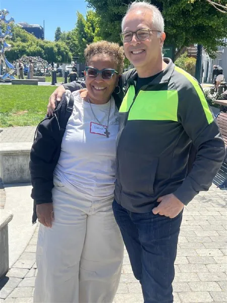 A smiling man and woman stand arm-in-arm outdoors on a sunny day. They appear relaxed, with greenery and people in the background.