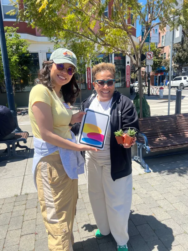 Two people smile outdoors on a sunny day, holding a colorful tablet and small potted plants. Trees and a bench are visible in the background.