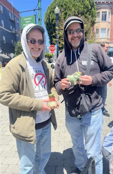 Two smiling men in hoodies and sunglasses stand outside on a sunny day holding succulent plants. They appear cheerful and relaxed, enjoying the moment.