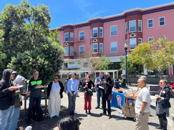 A group of people are gathered in a sunny outdoor plaza near pink buildings, engaging in conversation. Tables with refreshments are present. The mood is lively and social.